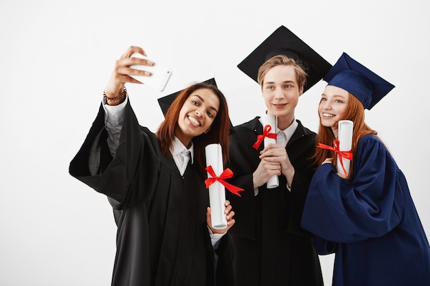 Cute friends graduates smiling holding diplomas making selfie.