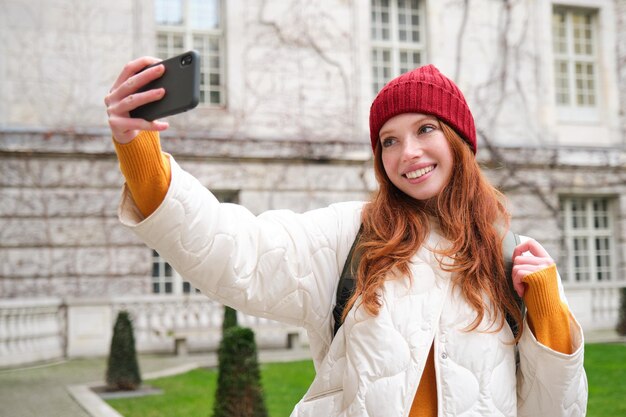 Cute ginger girl in red hat takes selfie during her tourist trip abroad young redhead woman makes a