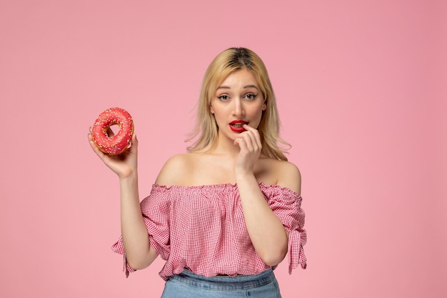 Cute girl adorable blonde chick with red lipstick in pink blouse biting finger holding a donut