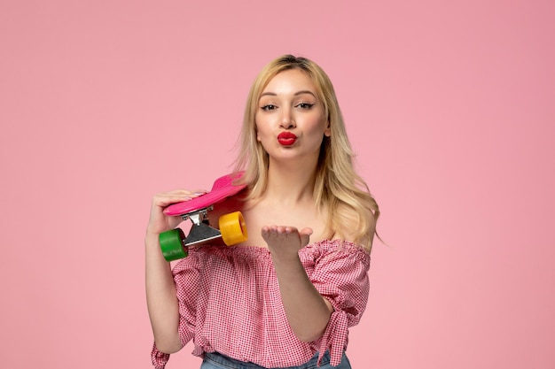 Cute girl adorable blonde chick with red lipstick in pink blouse sending kiss holding skateboard