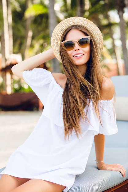 Cute girl in brown sunglasses and straw hat smiles charmingly.