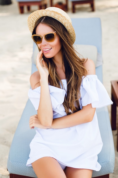 Cute girl in brown sunglasses and straw hat smiles widely and holds her hand near chin