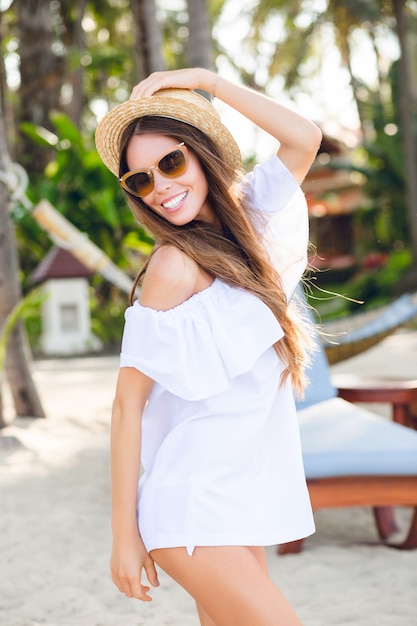 Cute girl in brown sunglasses and straw hat smiles widely and holds her hat with a hand