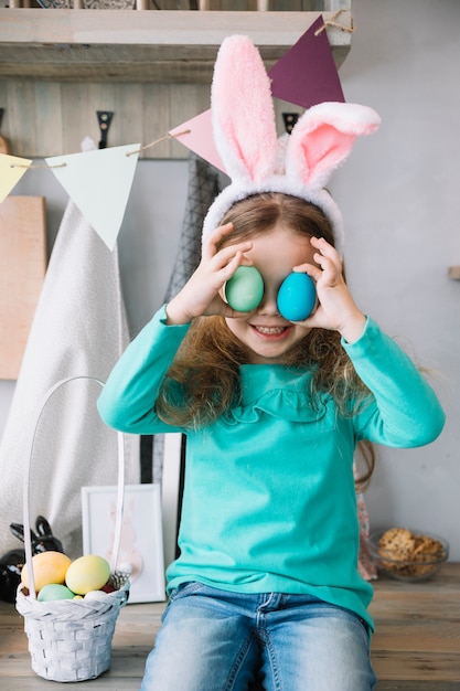 Cute girl in bunny ears holding colored eggs at eyes 