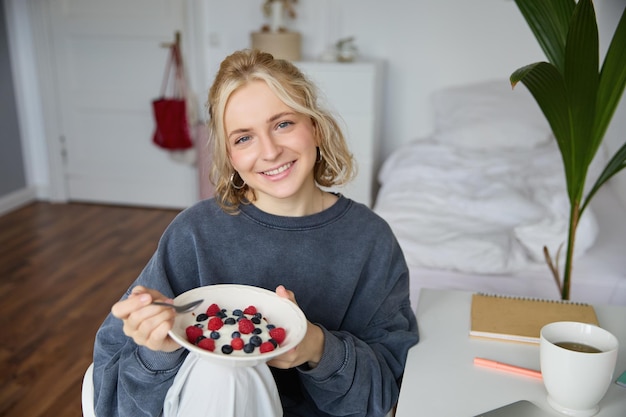 Cute girl eats breakfast and drinks tea in her room makes herself healthy lunch in bowl sits in