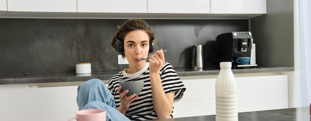 Free photo cute girl eats her breakfast holding bowl of cereals with milk drinks coffee and listens to music in