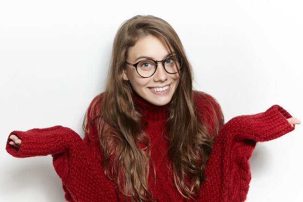 Free photo cute girl in eyewear being confused with unexpected question, smiling shyly, feeling embarrasses at lesson. female teenager wearing knitted sweater shrugging shoulders in confusion