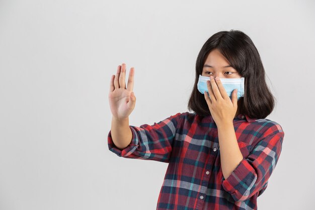 Cute girl is wearing mask and making stop hand from other prople on white wall.