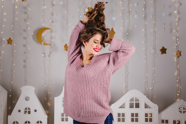 Free photo cute girl with bright lips in purple sweater plays hair and smiles against with luminous garlands and toy houses.