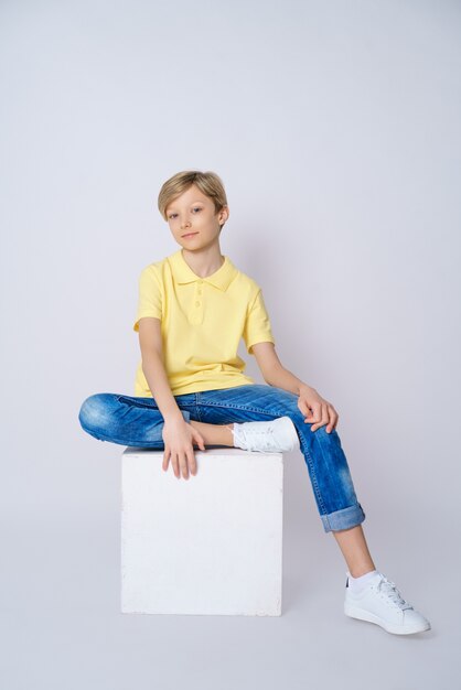 A cute guy in a yellow t shirt and blue jeans on a white background is sitting on a cube and posing