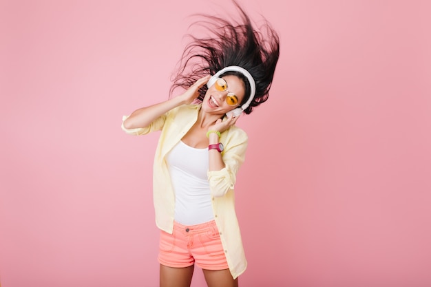 Free Photo cute hispanic girl with tanned skin wearing trendy bracelet and orange glasses listening music and dancing. indoor photo of attractive latin lady in yellow cotton jacket having fun.