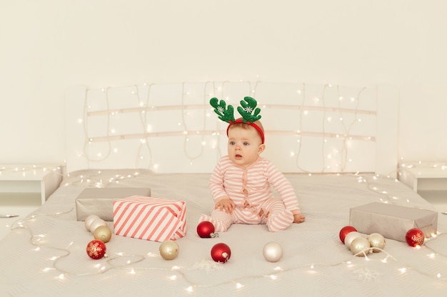 Cute infant girl sitting on Christmas decorations bed and wearing striped long sleeve baby sleeper and festive deer horns, looking at camera, celebrating new year.