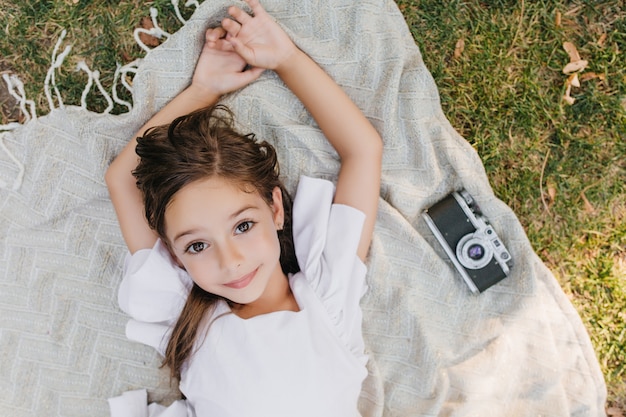 Free Photo cute lightly tanned girl with shiny beautiful eyes posing on blanket with camera during summer weekend. overhead portrait of brown-haired female kid lying on the grass and dreaming.