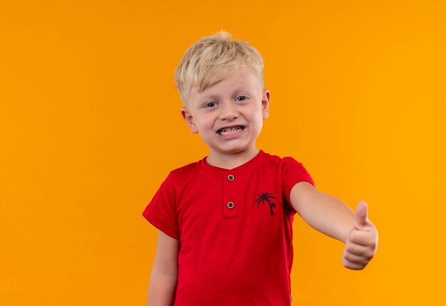 A cute little boy with blonde hair and blue eyes wearing red t-shirt showing thumbs up while looking on a yellow wall