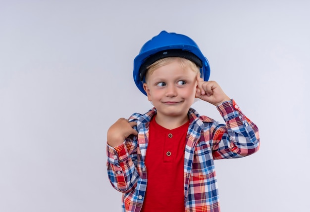 A cute little boy with blonde hair wearing checked shirt in blue helmet pointing at his head with index finger on a white wall