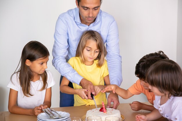 Cute little girl cutting birthday cake with help of father. Happy adorable children celebrating birthday together and waiting for dessert at dining room. Childhood, celebration and holiday concept