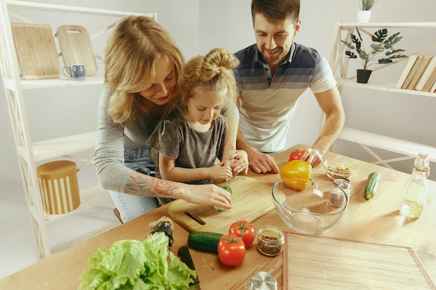 Cute little girl and her beautiful parents are cutting vegetables and smiling while making salad in kitchen at home. Family lifestyle concept