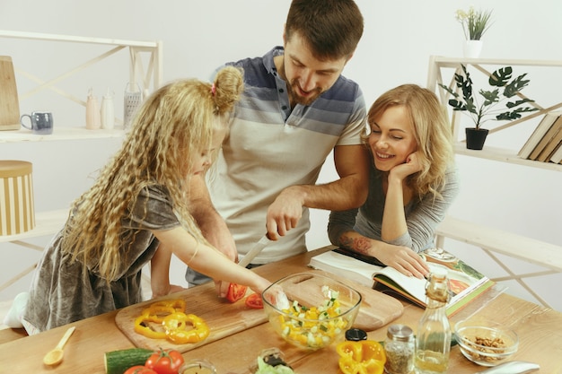 Free photo cute little girl and her beautiful parents are cutting vegetables and smiling while making salad in kitchen at home. family lifestyle concept