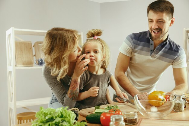 Cute little girl and her beautiful parents are cutting vegetables and smiling while making salad in kitchen at home. Family lifestyle concept