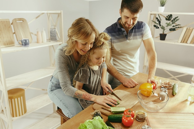 Free photo cute little girl and her beautiful parents are cutting vegetables and smiling while making salad in kitchen at home