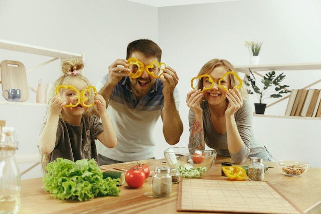 Free photo cute little girl and her beautiful parents are cutting vegetables and smiling while making salad in kitchen at home