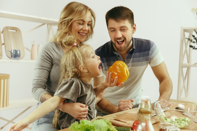 Free photo cute little girl and her beautiful parents are cutting vegetables and smiling while making salad in kitchen at home