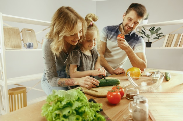 Cute little girl and her beautiful parents are cutting vegetables and smiling