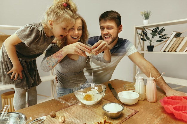 Cute little girl and her beautiful parents preparing the dough for the cake in kitchen at home