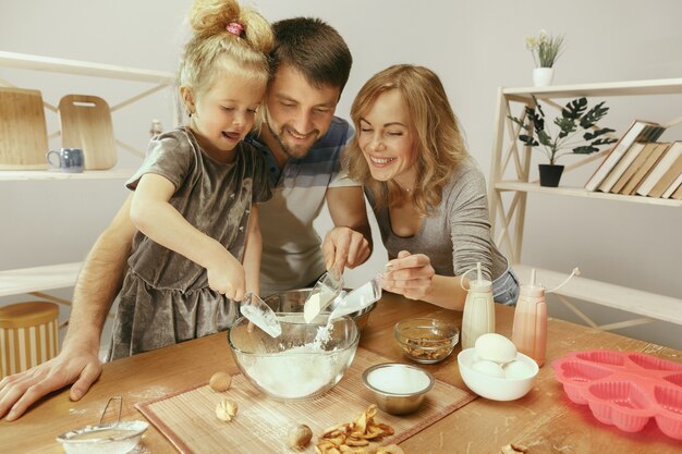 Cute little girl and her beautiful parents preparing the dough for the cake in kitchen at home