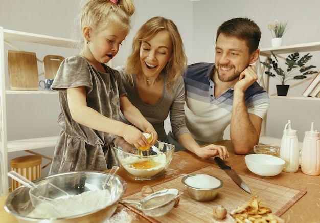 Cute little girl and her beautiful parents preparing the dough for the cake in kitchen at home