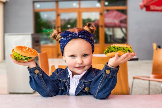 Cute little girl holding two burgers in hands in fast food cafe