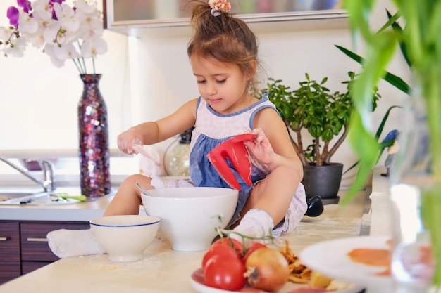 A cute little girl sits on a table in a kitchen and try to make diet porridge.