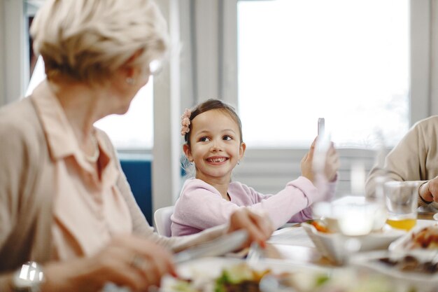 Cute little girl using smart phone and communicating with her grandmother during a meal at dining table