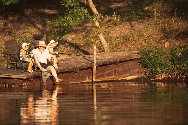 Cute little girls and their granddad are on fishing at the lake or river. Resting on pier near by water and forest in sunset time of summer day. Concept of family, recreation, childhood, nature.