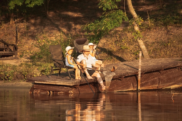 Cute little girls and their granddad are on fishing at the lake or river. Resting on pier near by water and forest in sunset time of summer day. Concept of family, recreation, childhood, nature.