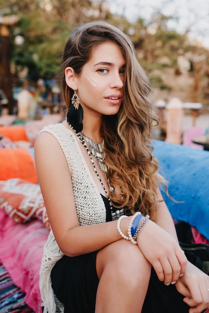 Cute long-haired girl in trendy bracelets and earrings posing outside while resting on the park party