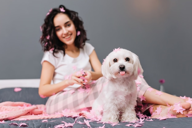 Free photo cute lovely little dog showing tongue on bed with pretty young woman. chilling at home with domestic animals, funny moments