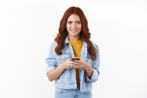 Free photo cute modern young redhead girl with freckles in denim jacket, hold smartphone and smiling delighted, smm freelancer work remote, contact customers via mobile phone, white wall