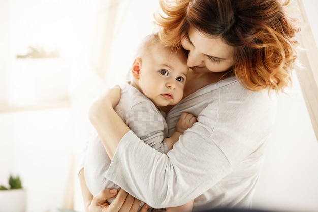 Cute newborn kid frightenedly looks aside while good-looking young mother gently huggs baby expressing her love and support. Family concept.