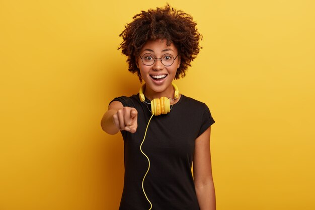 Cute positive dark skinned girl wears balck t shirt, round glasses, has yellow headpohones connected to some gadget, being real meloman