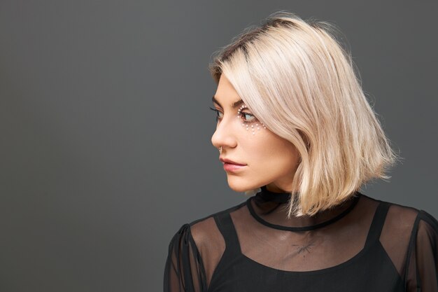 Cute positive young Caucasian female wearing trendy black top and transparent dress having dreamy pensive facial expression, making plans for Christmas holidays, looking away and smiling joyfully