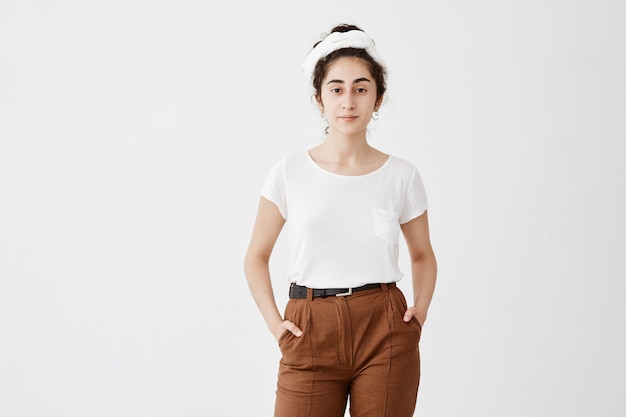 Free Photo cute pretty teenage girl wears loose white t-shirt and brown trousers, holds her hands in pocket, feels enjoyment. pleasant-looking girl posing against white wall