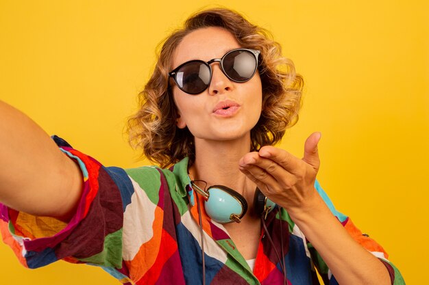 Cute pretty woman with earphones send kiss to camera and making self portrait over yellow wall