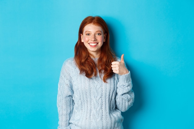 Free photo cute redhead girl in sweater showing thumb up, like and agree, smiling pleased, standing against blue background