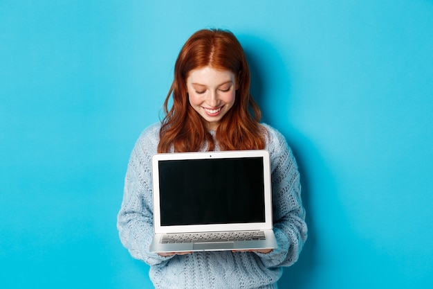 Cute redhead woman in sweater, showing and looking at laptop screen with pleased smile, demonstrating something online, standing over blue background