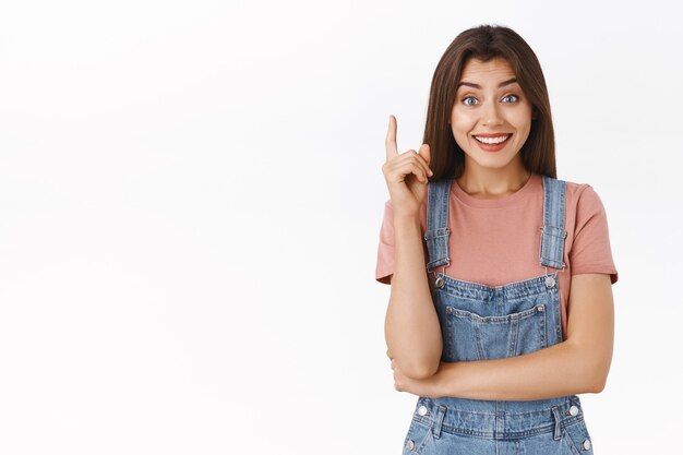 Cute relieved girl finally got excellent idea, sharing her plan or suggestion, smiling happy and silly looking camera, raise one finger in eureka gesture, got solution, standing white background