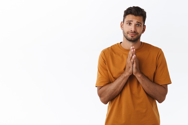 Cute silly attractive hipster man in brown tshirt making promise smiling with angelic innocent smile press hands together over chest praying or asking help need something white background