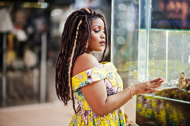 Cute small height african american girl with dreadlocks wear at coloured yellow dress looking aquarium at shopping center