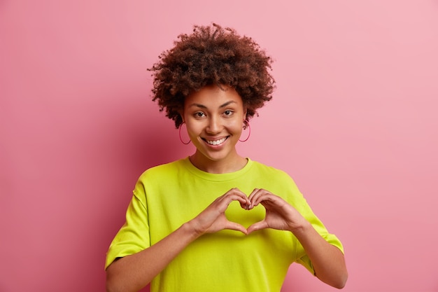 Free photo cute smiling afro american woman makes i love you gesture confesses in love expresses sympathy shows heart sign dressed in casual clothes isolated over pink wall