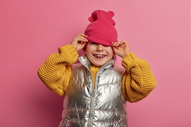 Free photo cute smiling girl looks from under hat, dressed casually, has toothy smile, going crazy, shows two adult teeth, enjoys spare time for playing with friends, isolated on pink wall. happy childhood